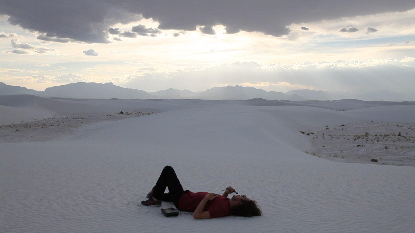 Color photograph of a man lying in the middle of a desert speaking into a microphone attached to a recorder