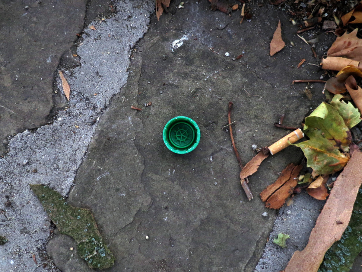 A photograph of a green plastic bottle cap on the street, next to a pile of leaves.
