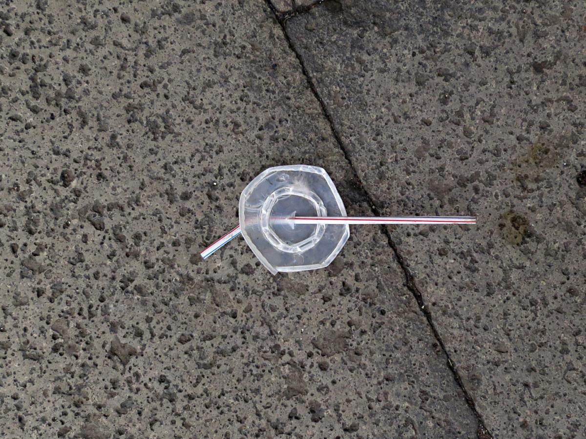 A photograph of a disposable drink lid & striped plastic straw on the sidewalk