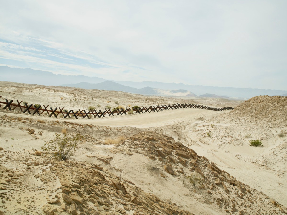 Color photograph of a metal fence made of X-shaped crossbars running through a hilly desert landscape