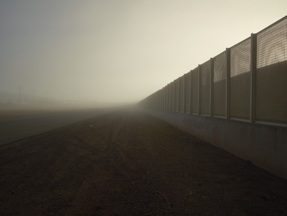 Color photograph of a tall metal and concrete fence receding into the misty horizon on the right