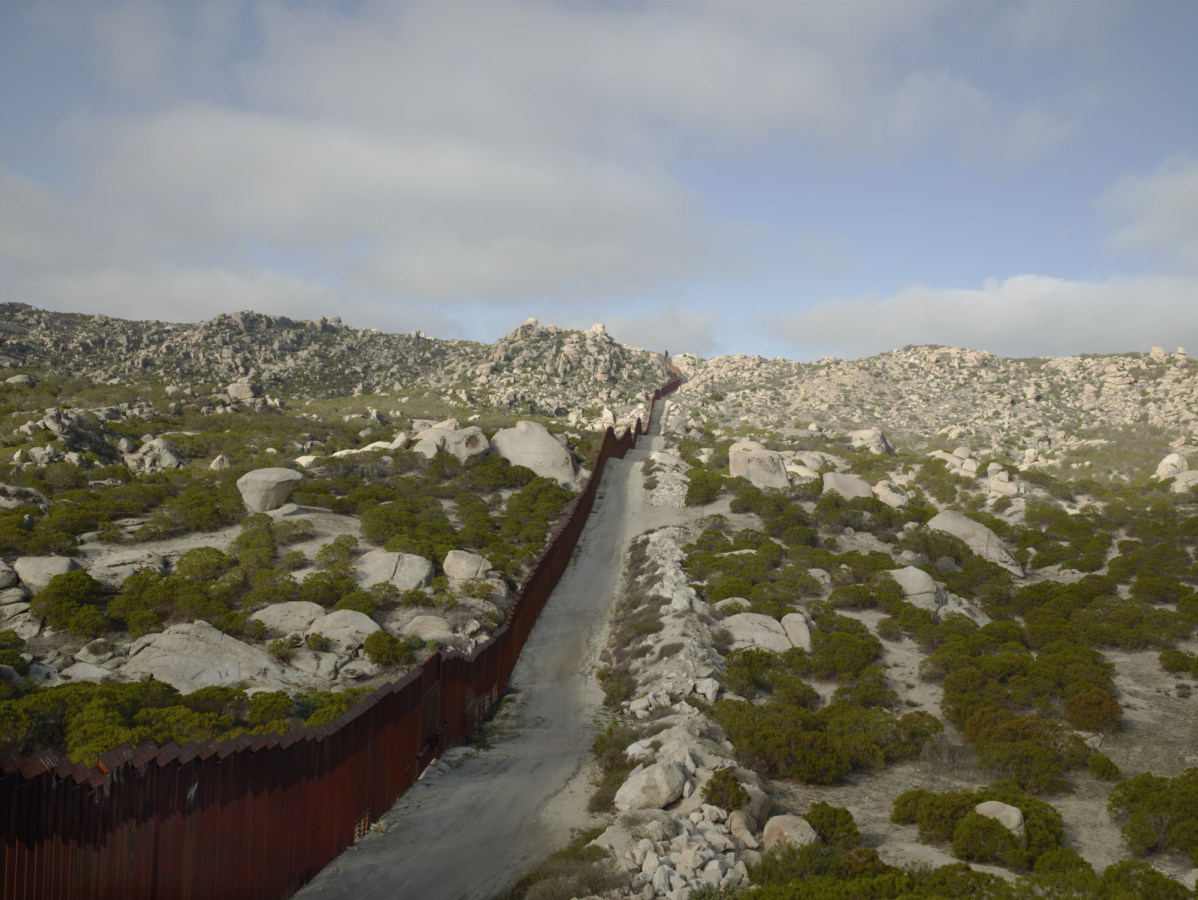 Color photograph of a metal fence running over a landscape of rocky hills and low green bushes into the horizon