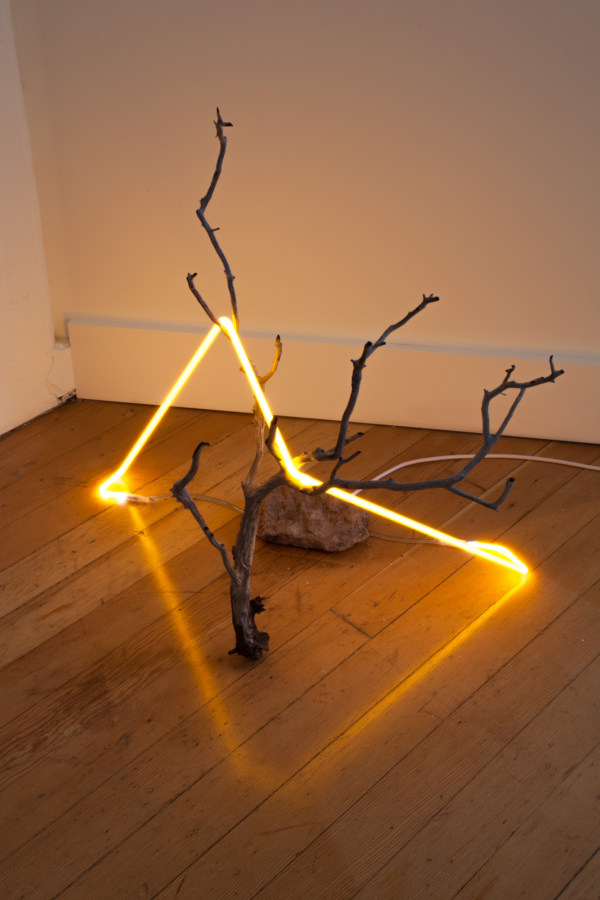 Color image of a sculpture with a yellow neon light atop a branch wired through a rock on gallery floor