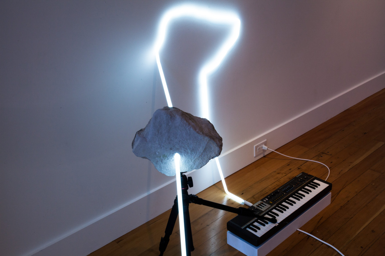 Color image detail of artwork with white neon light poking through a rock attached to small keyboard