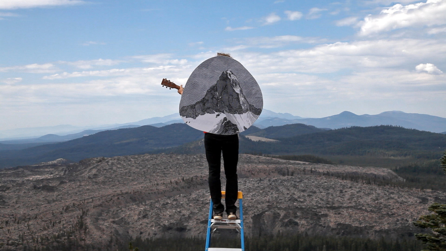 Video still of a person on a step stool holding a guitar in a mountainous landscape with a circular illustration of a mountain overlaid on top of him