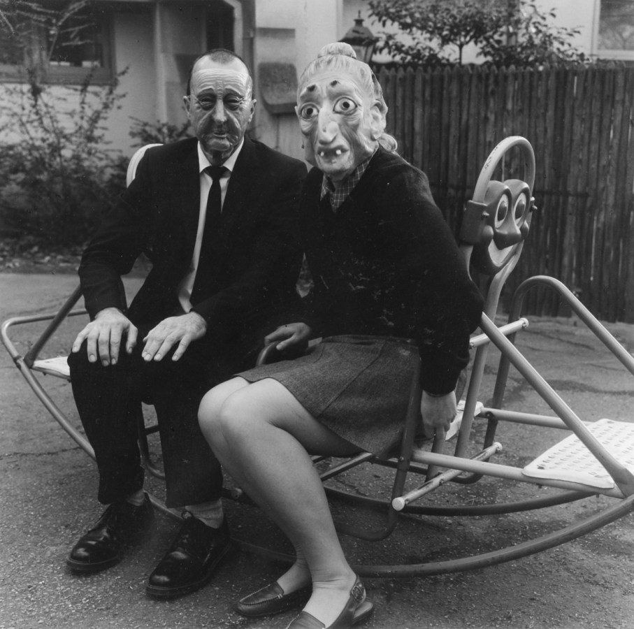 Black and white photograph of two people in Halloween masks seated on a see saw.