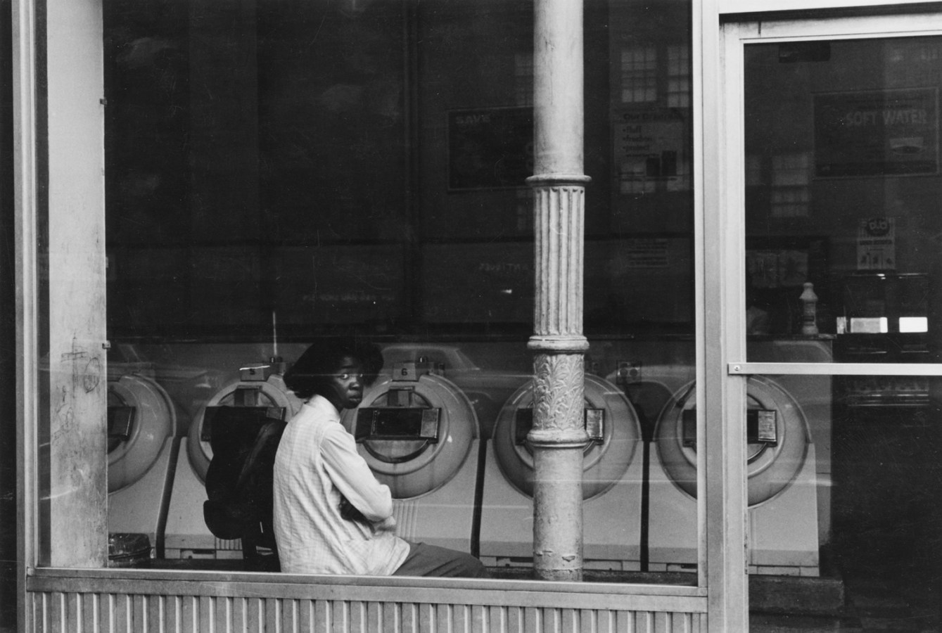 Black and white photograph of an African American woman seated in the window of a laundromat.