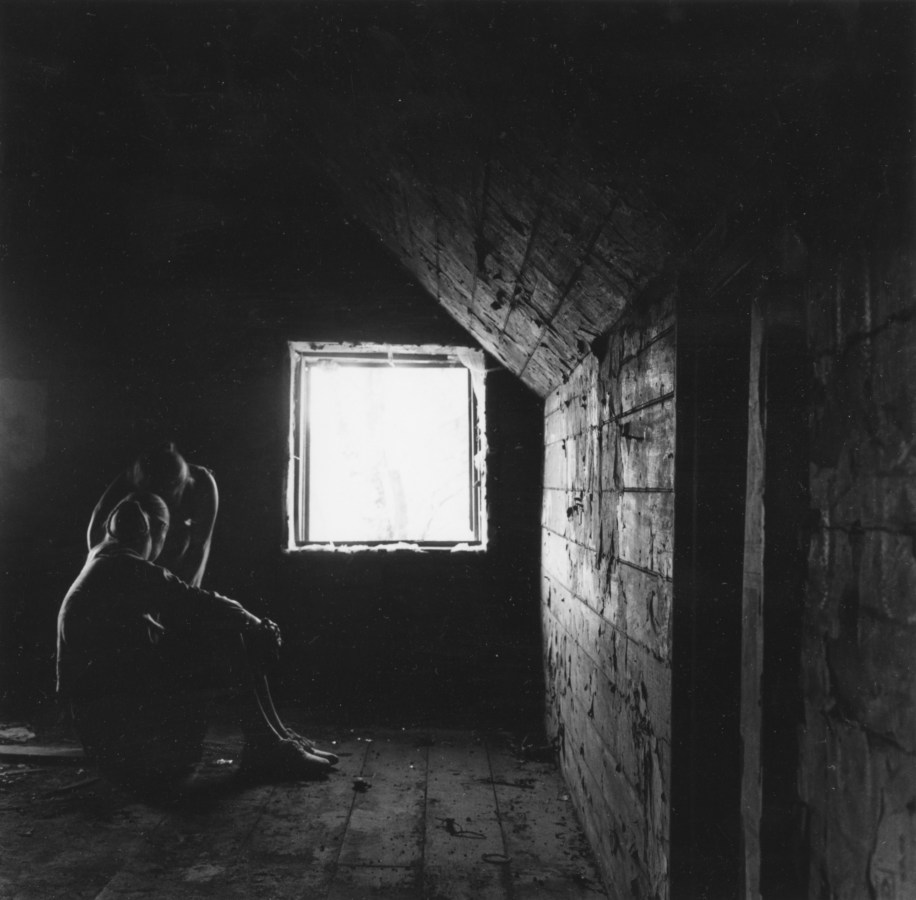 Black-and-white photograph of two figures seated in an attic room next to a square window