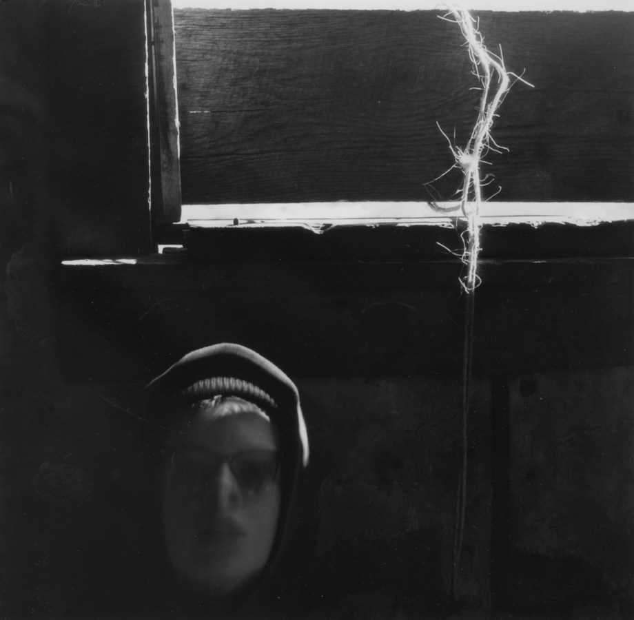 Black-and-white photograph of a person's slightly out-of-focus and shadowed face beneath a strand of twine coming through a window