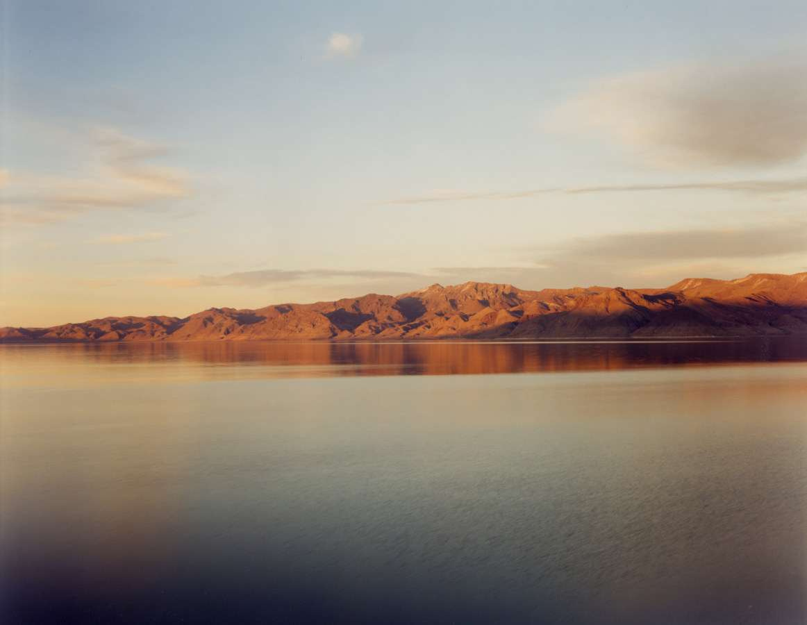 Color photograph of a low mountain range reflected in still water at sunset
