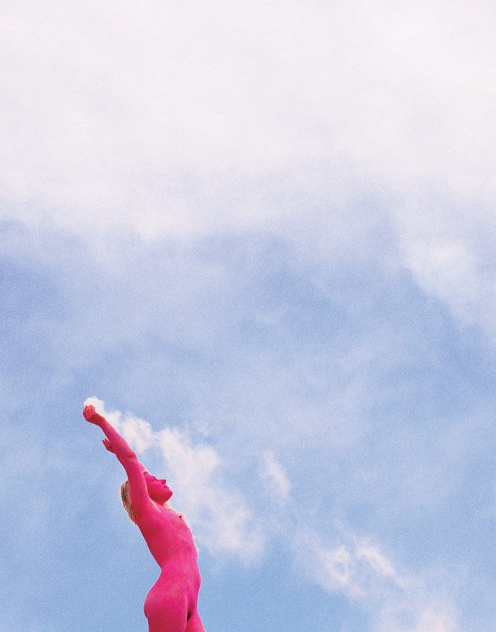 Color photograph of nude figure painted fully in pink holding a smoke flare with cloudy sky behind the firgure