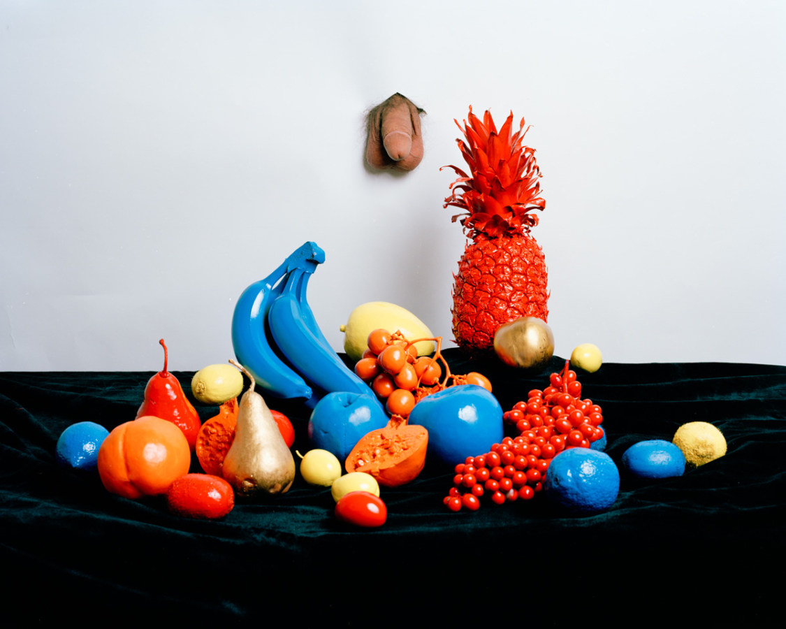 Color photograph of fruit still life with fruits painted in primary colors in front of male genitals poking through white background