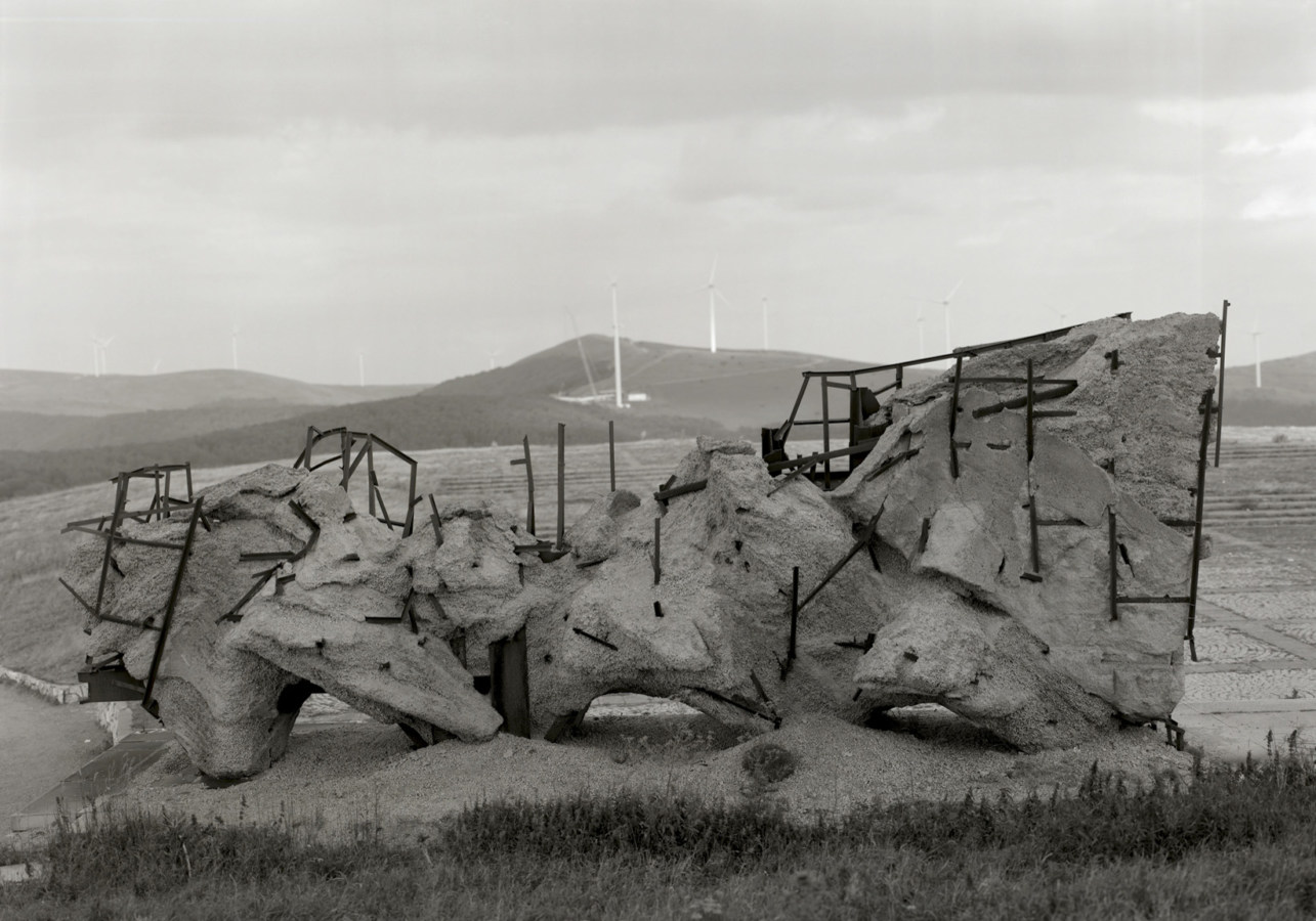 Black and white horizontal photograph of a concrete-and-metal armature in a hilly landscape.