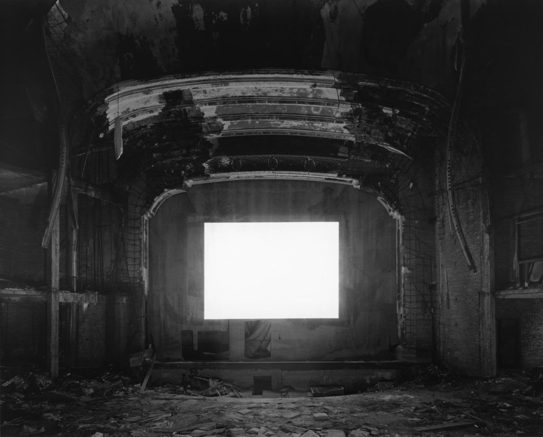 Black-and-white photograph of an empty abandoned theater with a glowing blank white screen