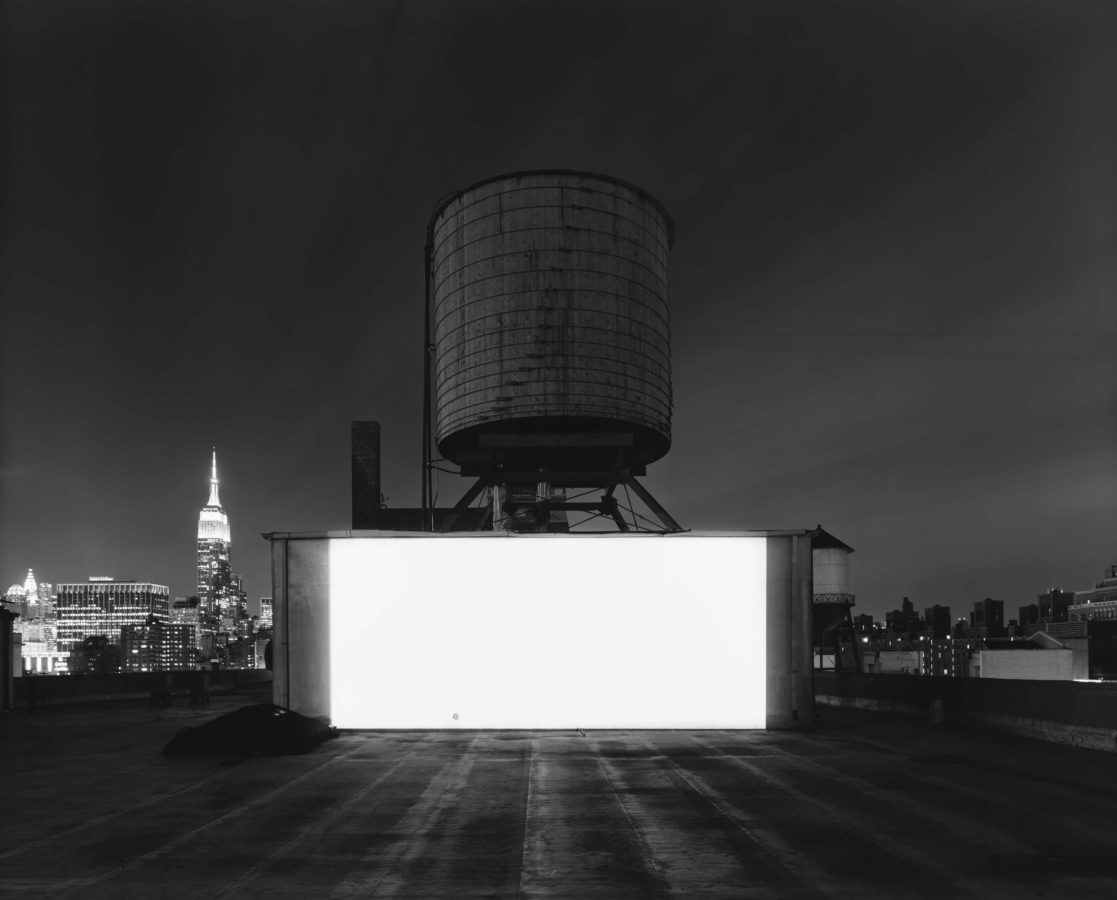 Black-and-white photograph of a glowing blank white screen against a water tower and city night skyline