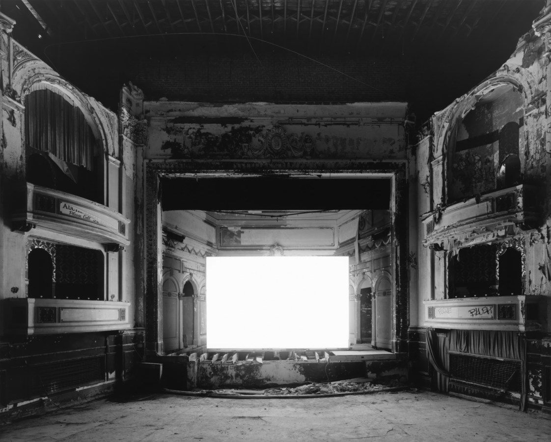 Black-and-white photograph of an empty abandoned theater with a glowing blank white screen