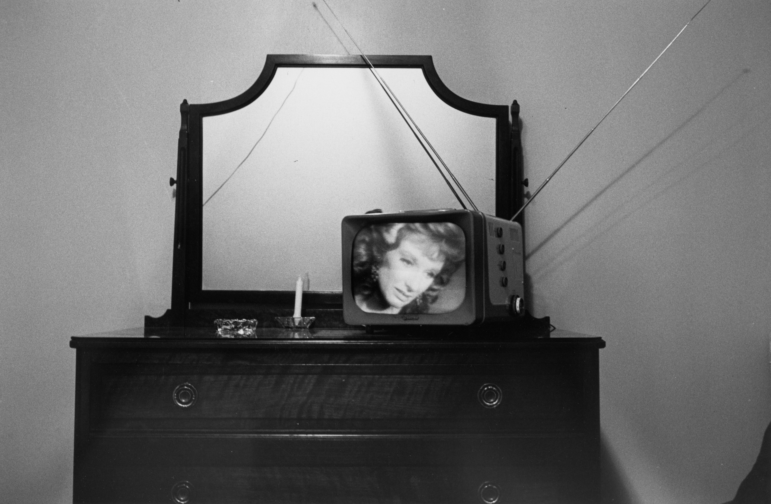Black-and-white photograph of a television with a woman's face onscreen on top of a bureau