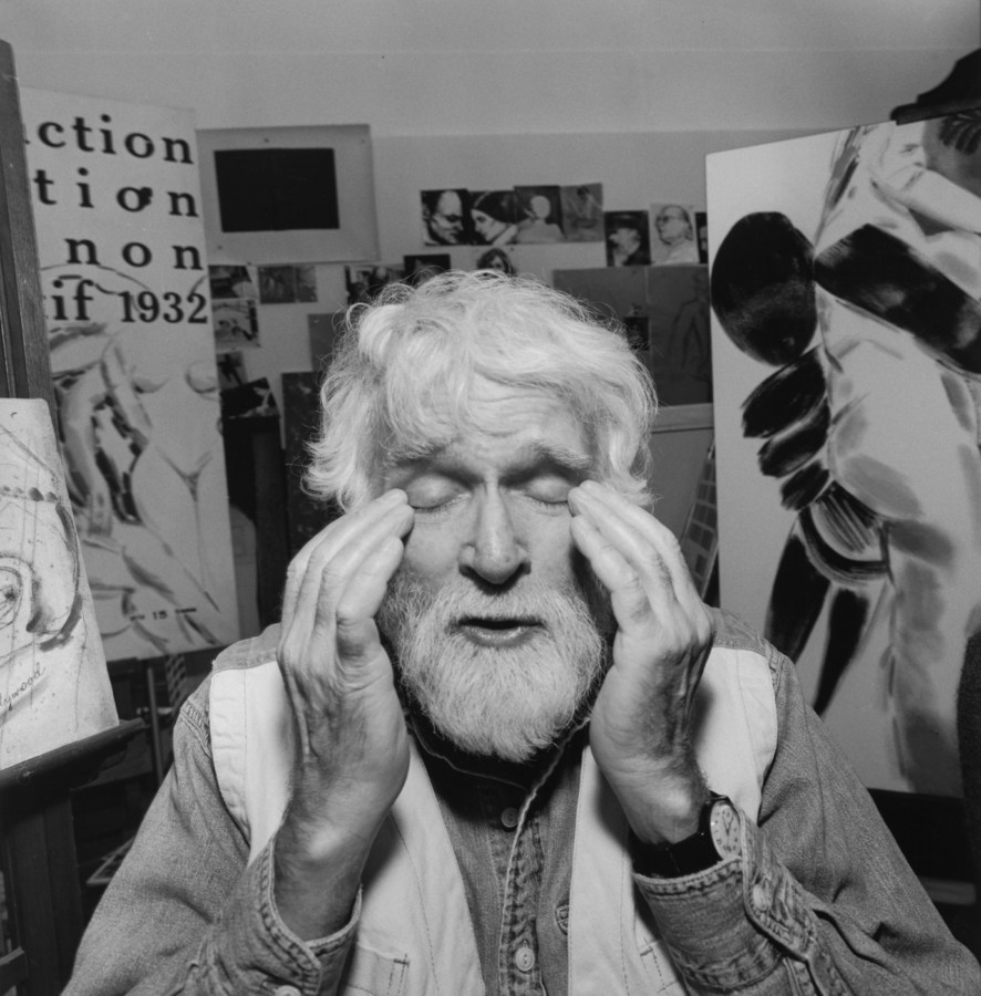 Black-and-white photograph of a man rubbing his eyes with paintings in the background