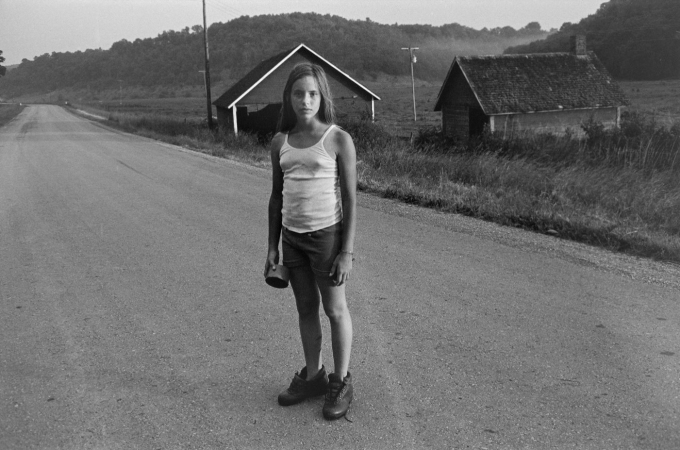 Black-and-white photograph of a girl standing in a dirt road with her head framed by a building in the background