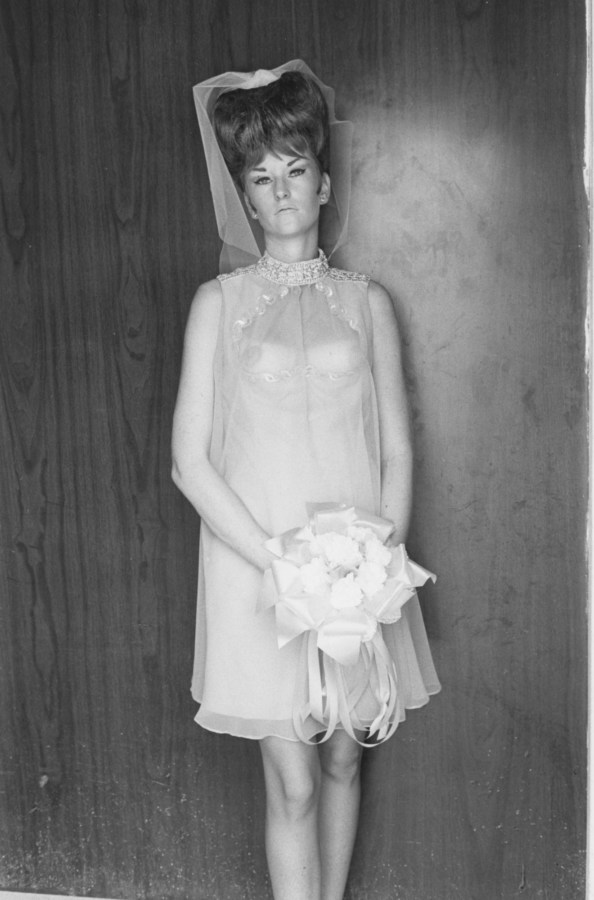 A black and white photograph of a woman wearing a see through bridesmaid dress and holding a bouquet in front of a wood paneled wall