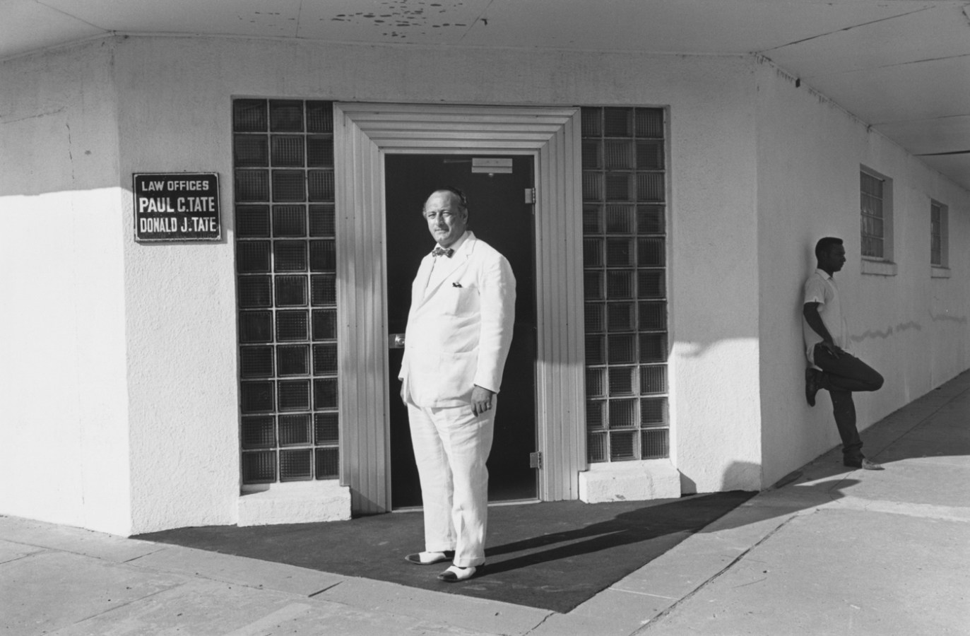 Black-and-white photograph of a man in a white suit standing in front of a doorway and another man to his right in the background