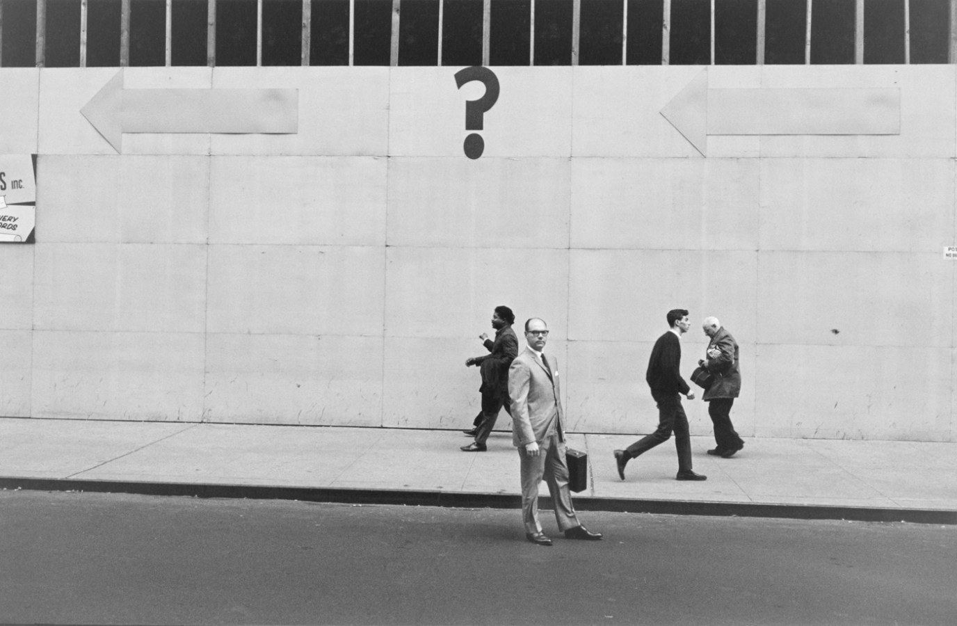 Black-and-white photograph of a man in a suit standing in the street in front of a. building with a question mark painted on it