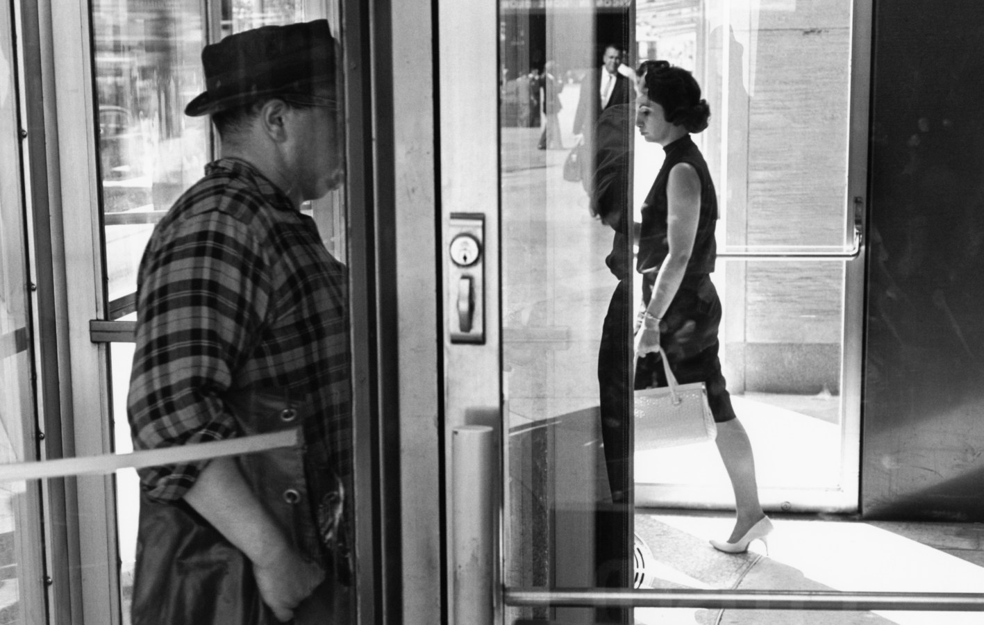 Black-and-white photograph of a man and woman entering and exiting a building with their faces obscured by doors
