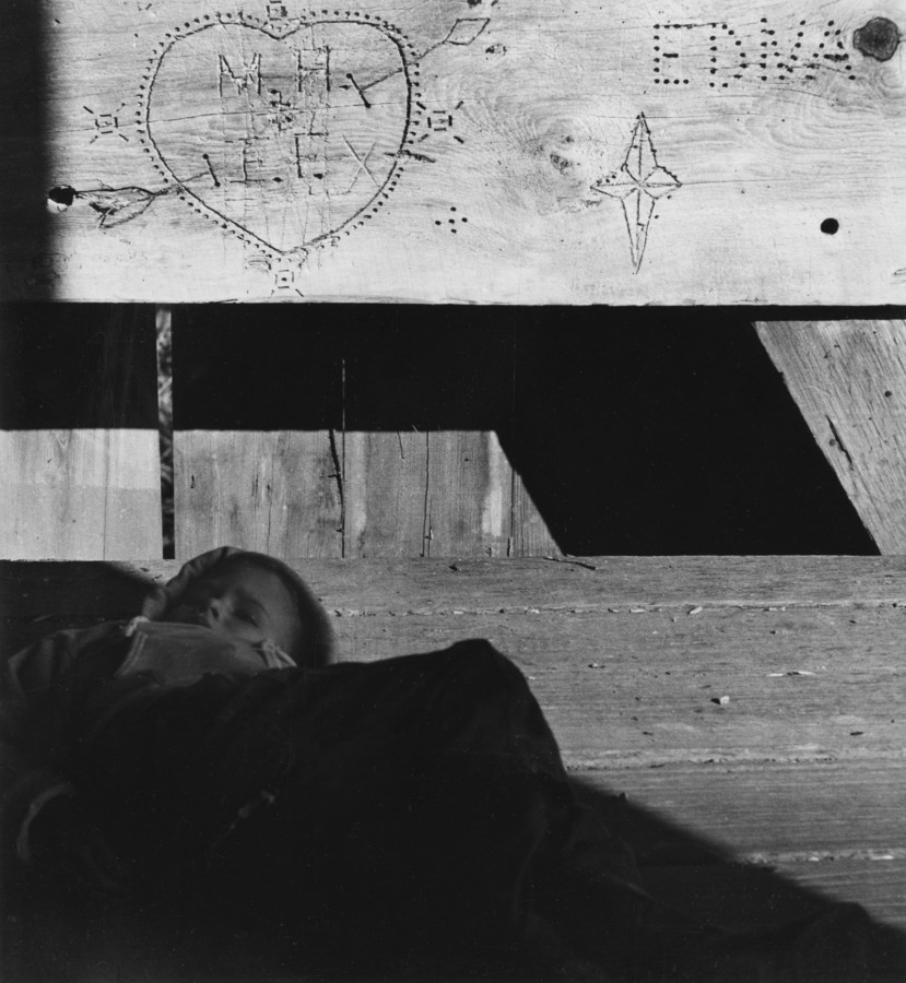 Black and white photograph of young boy with eyes closed; above him is a wood beam with various incised inscriptions.