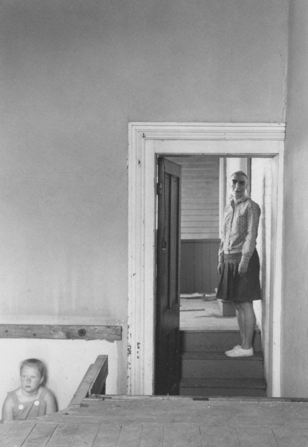 Black-and-white photograph of a woman in a mask standing in a doorway with a child in overalls on the lower left