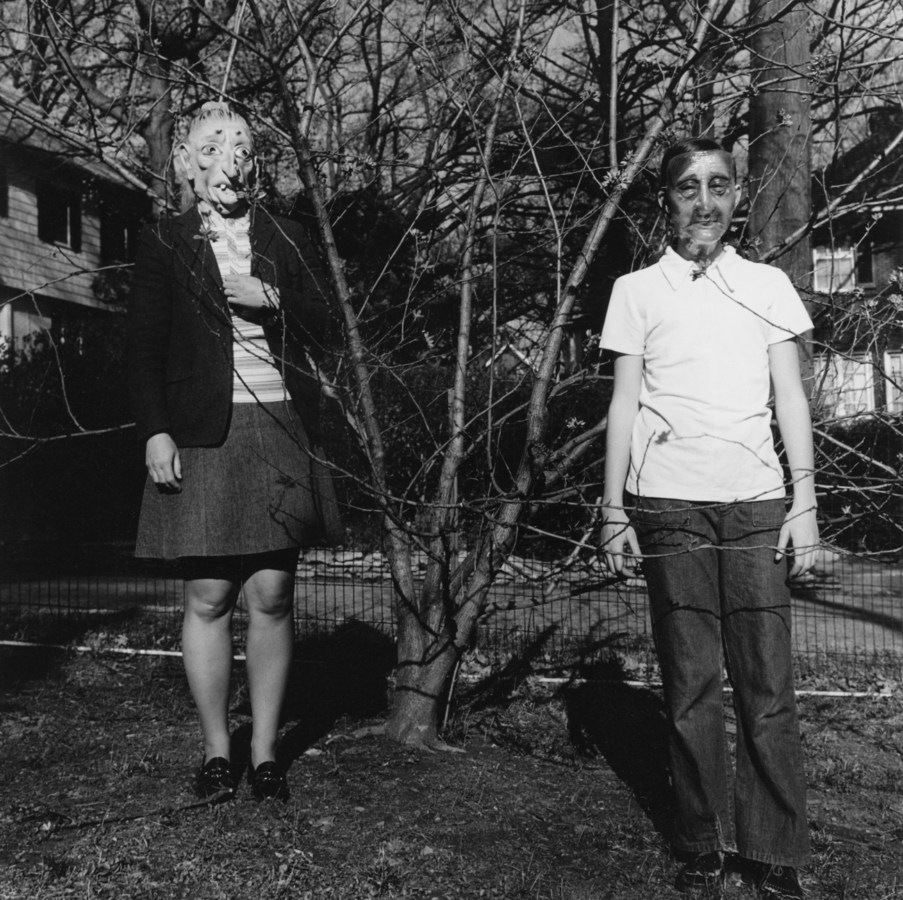 Black and white photograph of two people in Halloween masks standing on either side of a leafless tree.