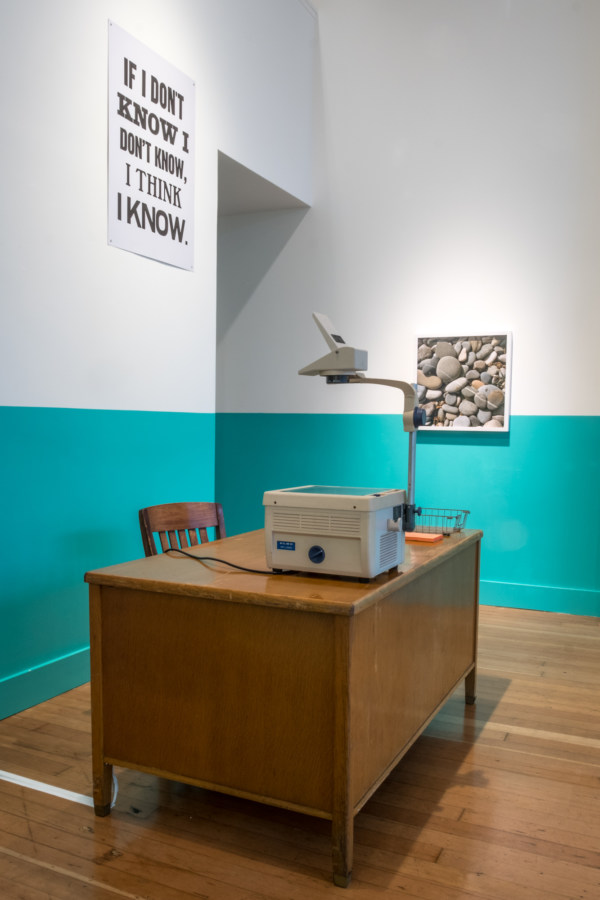 Color image of framed color photographs on white and teal gallery walls with desk and overhead projector