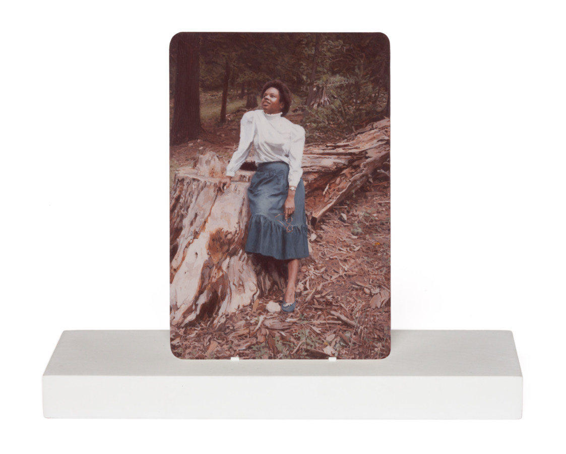 Color image of photorealistic painting of photograph depicting a young woman looking upward at sky while leaning on tree stump on white stand