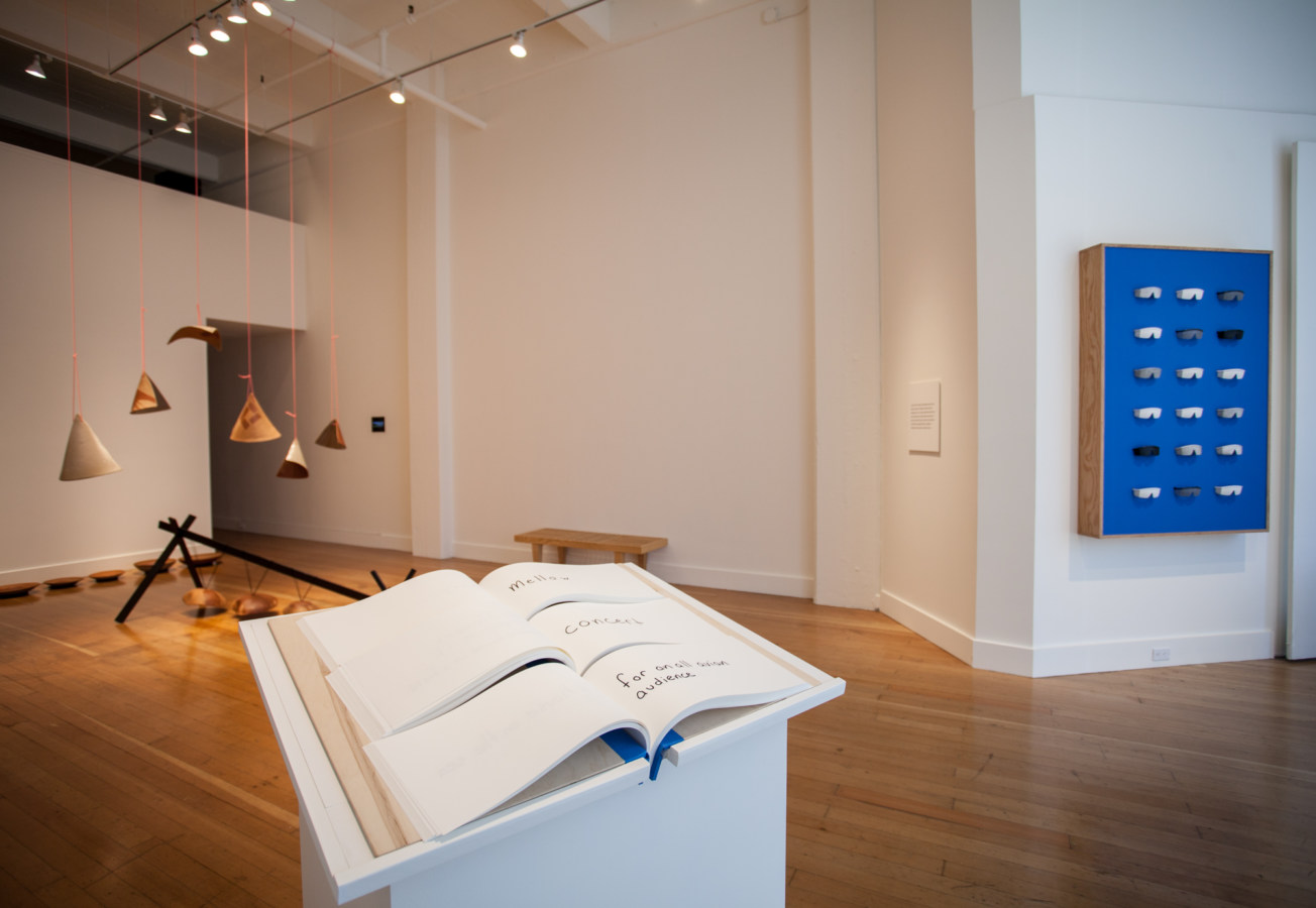 Color image of white walled gallery exhibiting sculptural sound artworks and book on white pedestal