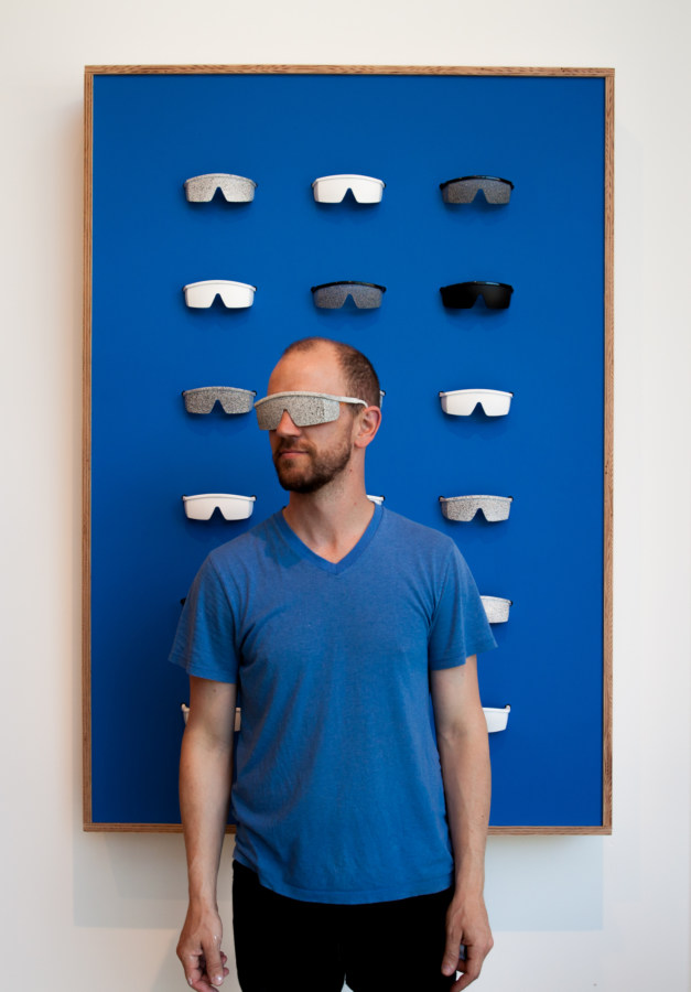 Color image of man in blue shirt with sensory deprivation glasses on in front of blue artwork with removable sensory deprivation glasses