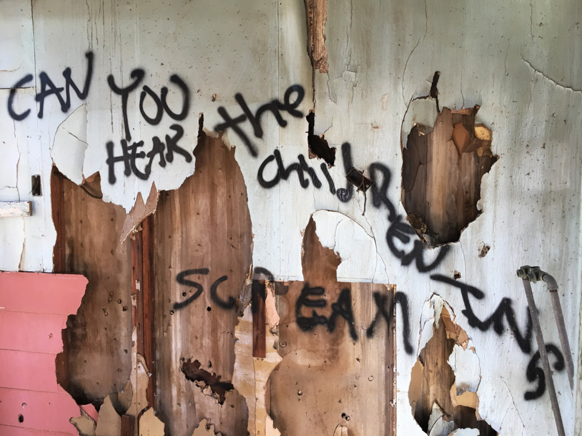 Color photograph of black spray-painted writing on a peeling and partly-demolished white and wooden wall