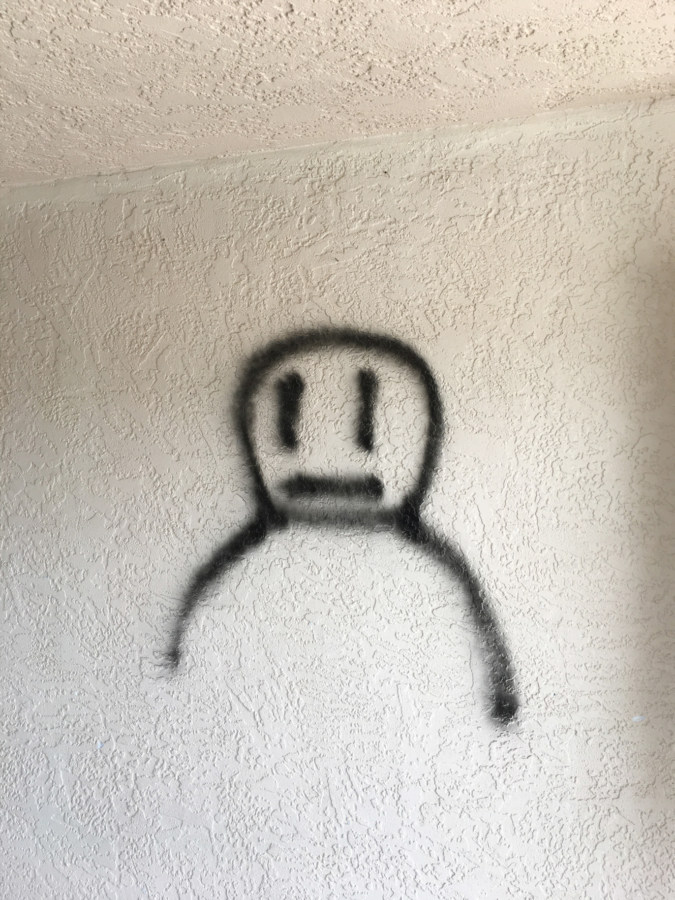 Color photograph of a simple black spray-painted face and shoulders on a white wall