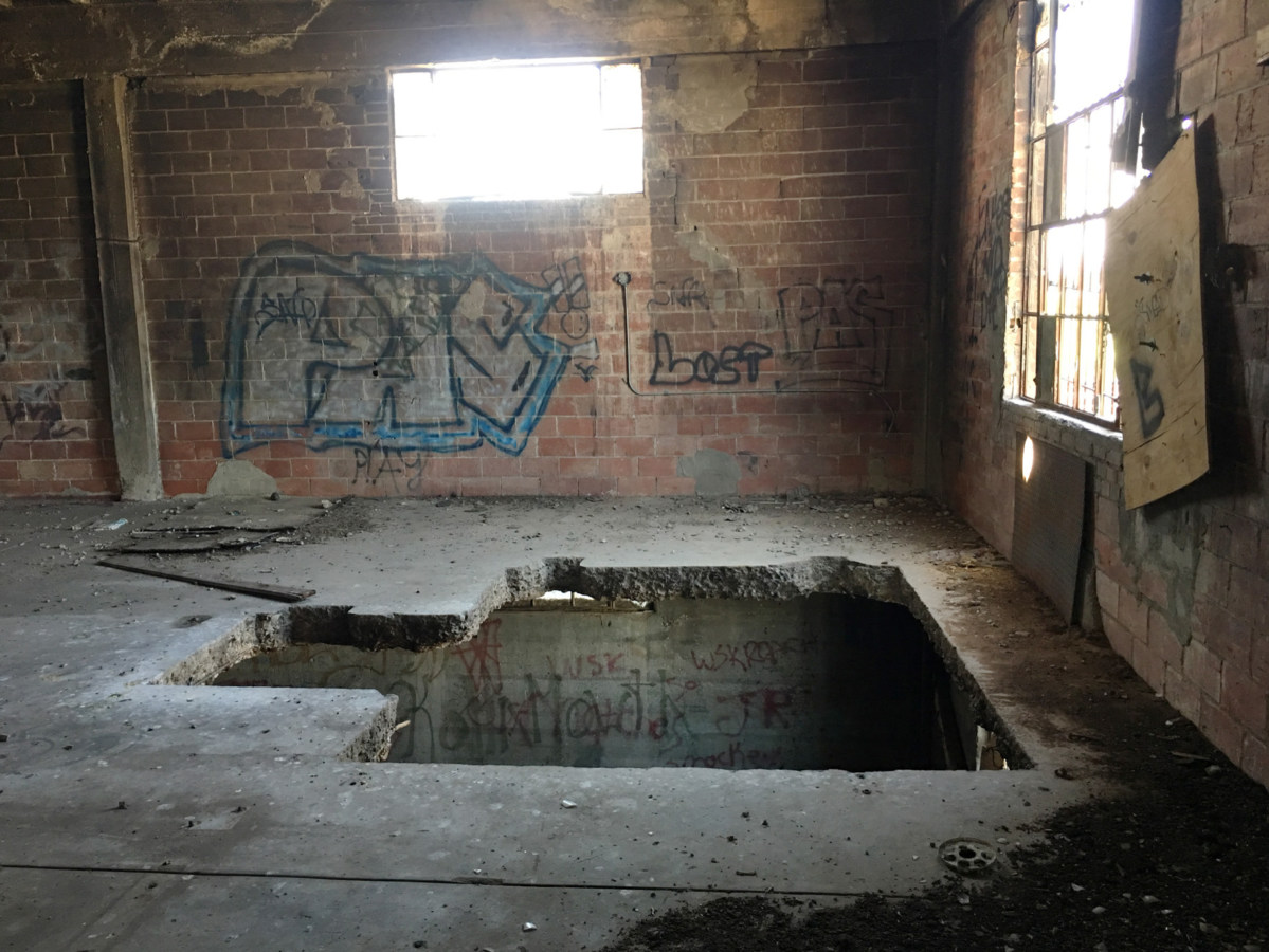 Color photograph of the spray-painted interior of an abandoned brick building with a hole in the floor leading to the lower level
