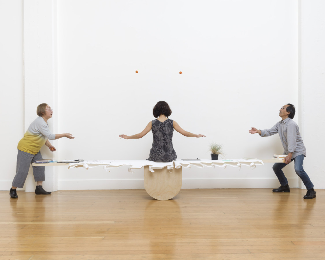 Color photograph of three performers in white walled gallery two are on either end of a seesaw tossing mandarins while the other sits in the center back to camera arms outstretched