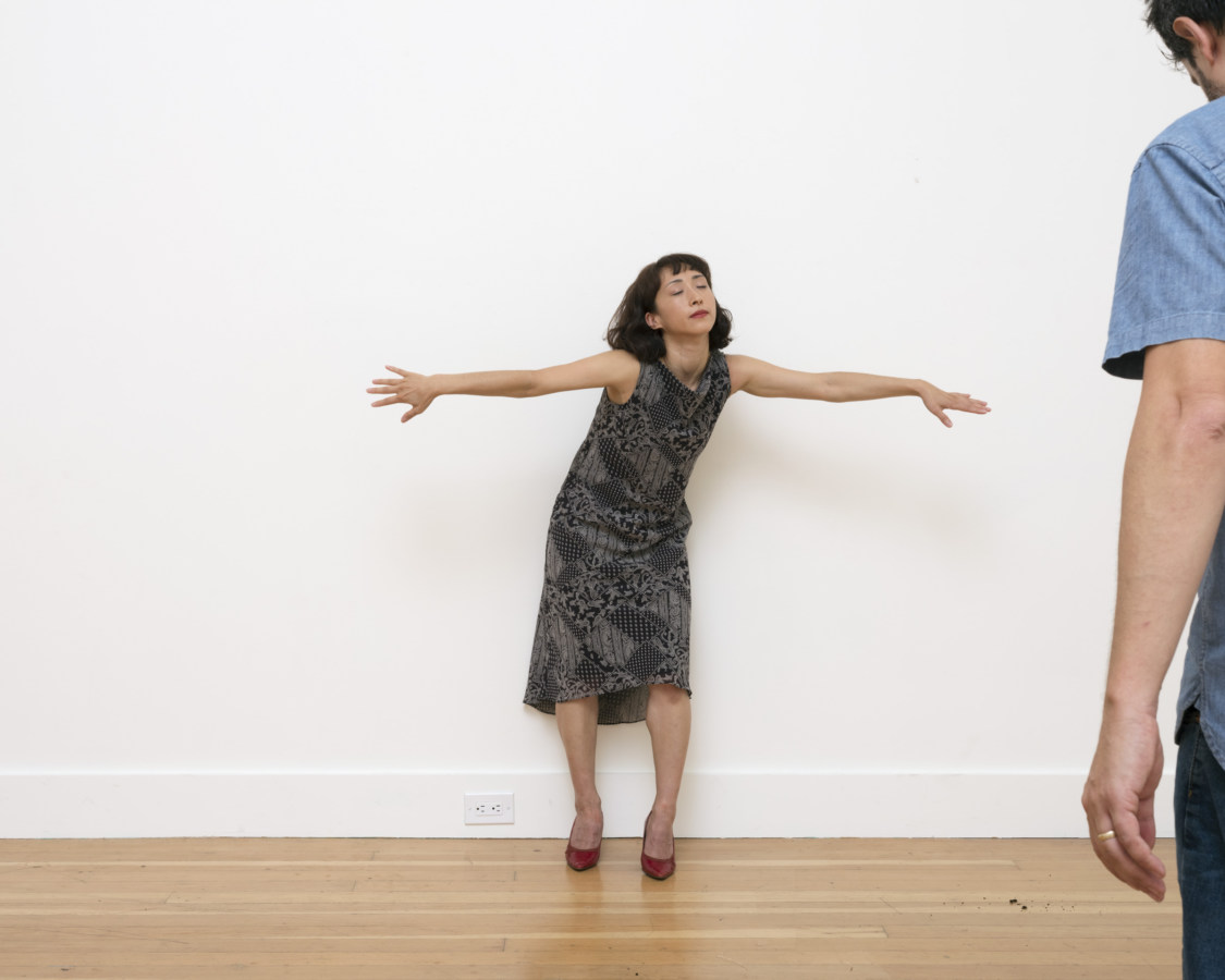 Color photograph of women performer in grey dress and red shoes with arms out stretched and eyes closed in white gallery