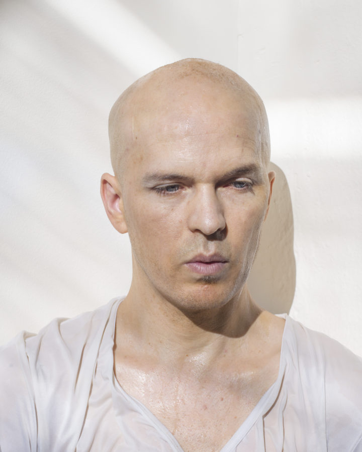 Color photograph of a bald man drenched in water in front of white gallery wall