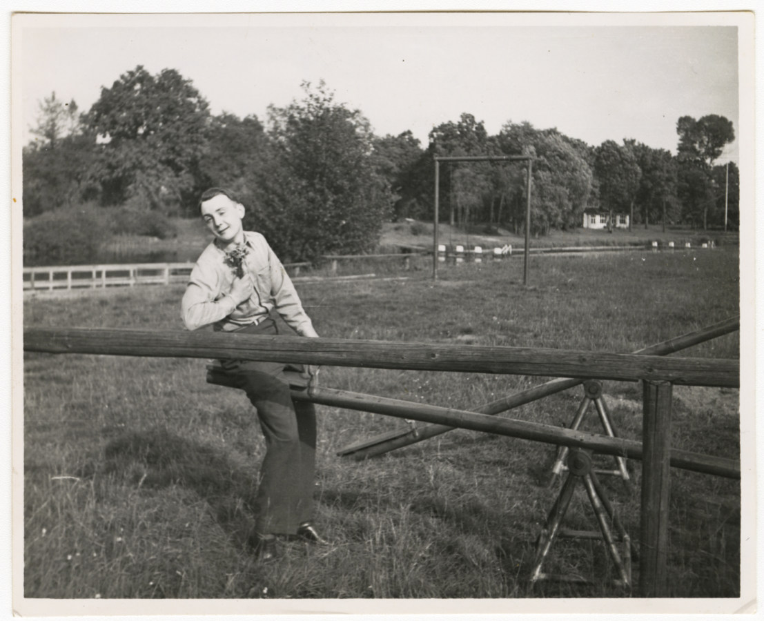 Black and white photograph of young man holding flowers while on wooden seesaw in a grassy field