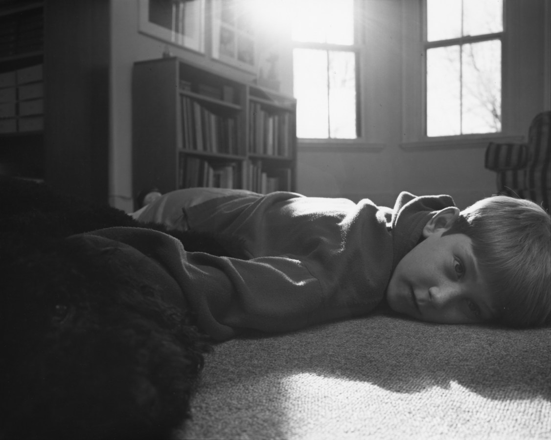 Black-and-white photograph of a boy lying on the floor of a sunny room with his face turned to the viewer