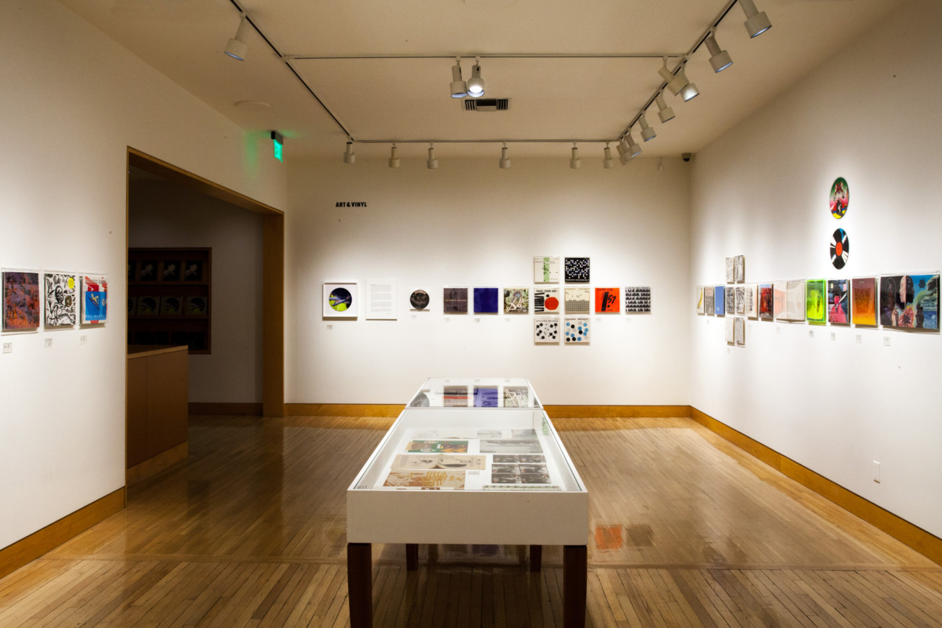 Color image of vinyl records and record sleeves inside white vitrine and on white gallery walls
