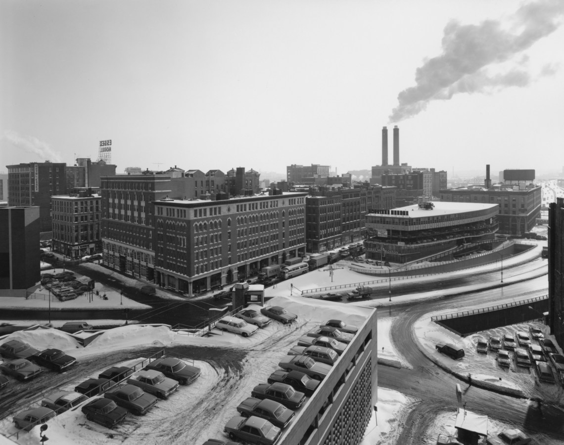 Black-and-white photograph of a city intersection in winter with a rooftop garage and smokestacks