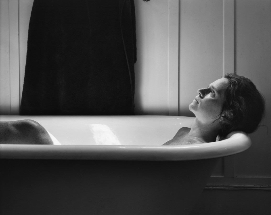 Black-and-white photograph of a woman reclining in a white bathtub looking up