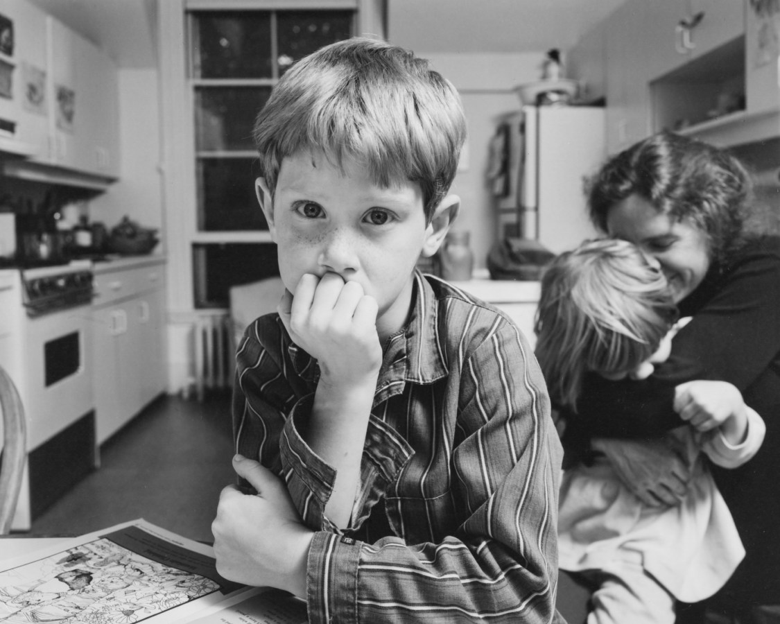 Black-and-white photograph of a boy seated in a kitchen with his chin in his hand with a woman and child behind him