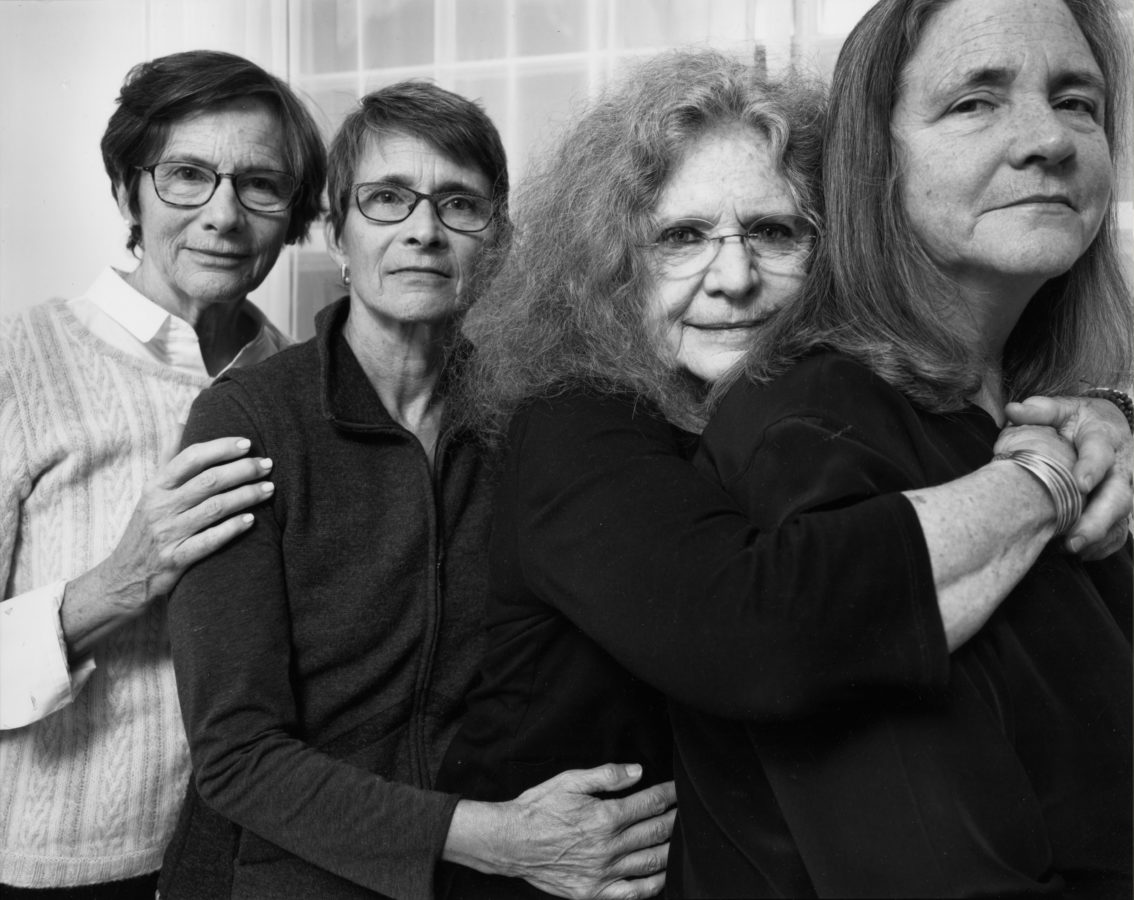 Black-and-white photographic portrait of four older women with their arms around each other standing in front of a curtained window
