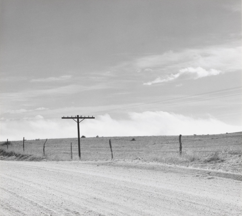 A black and white photograph of a road, with sky in the first half of the photograph. A telephone pole is in the center left