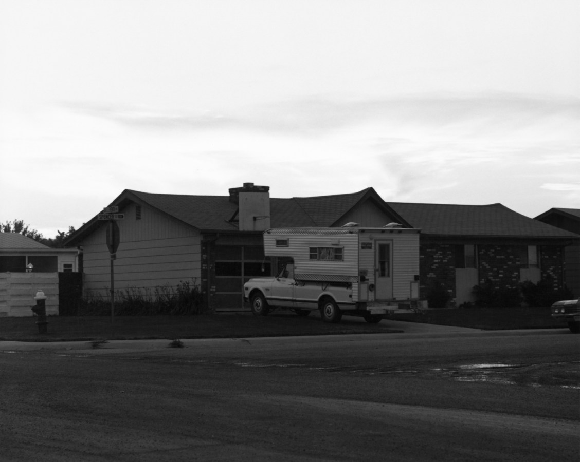 A black and white photograph of a pickup truck camper parked in the driveway of a suburban house.