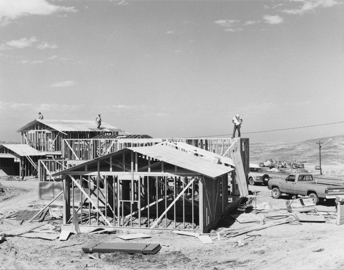 A black and white photograph of three construction workers on the roofs of unfinished houses.